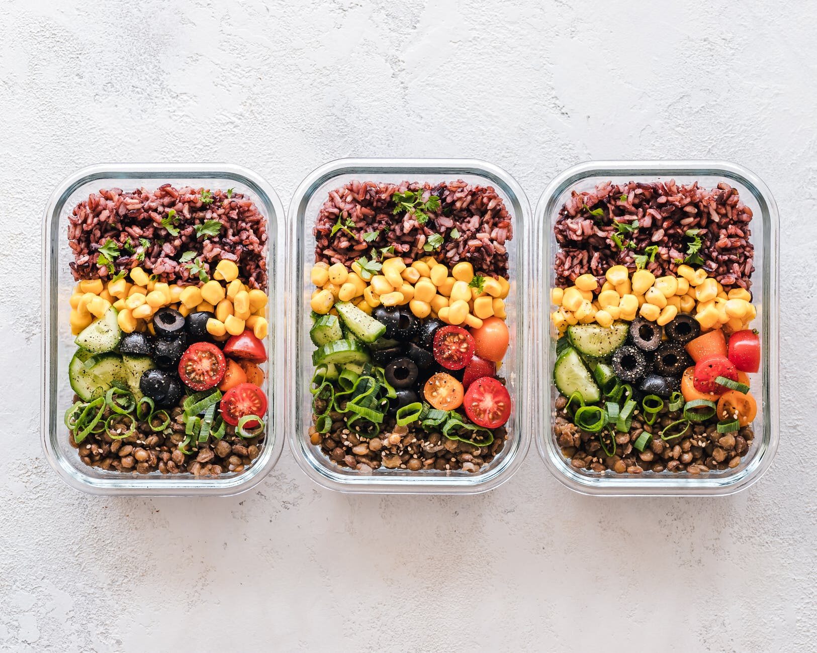 Three glass containers filled with colorful prepared salads and toppings on a white table